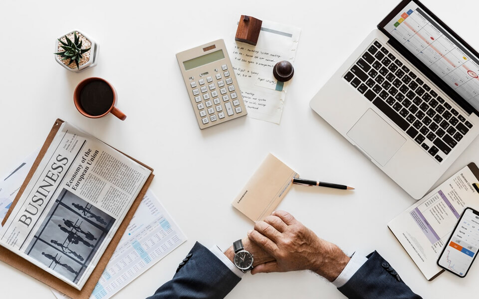 A professional businessman's hands rest on a desk, surrounded by a laptop, newspaper, calculator, and papers.